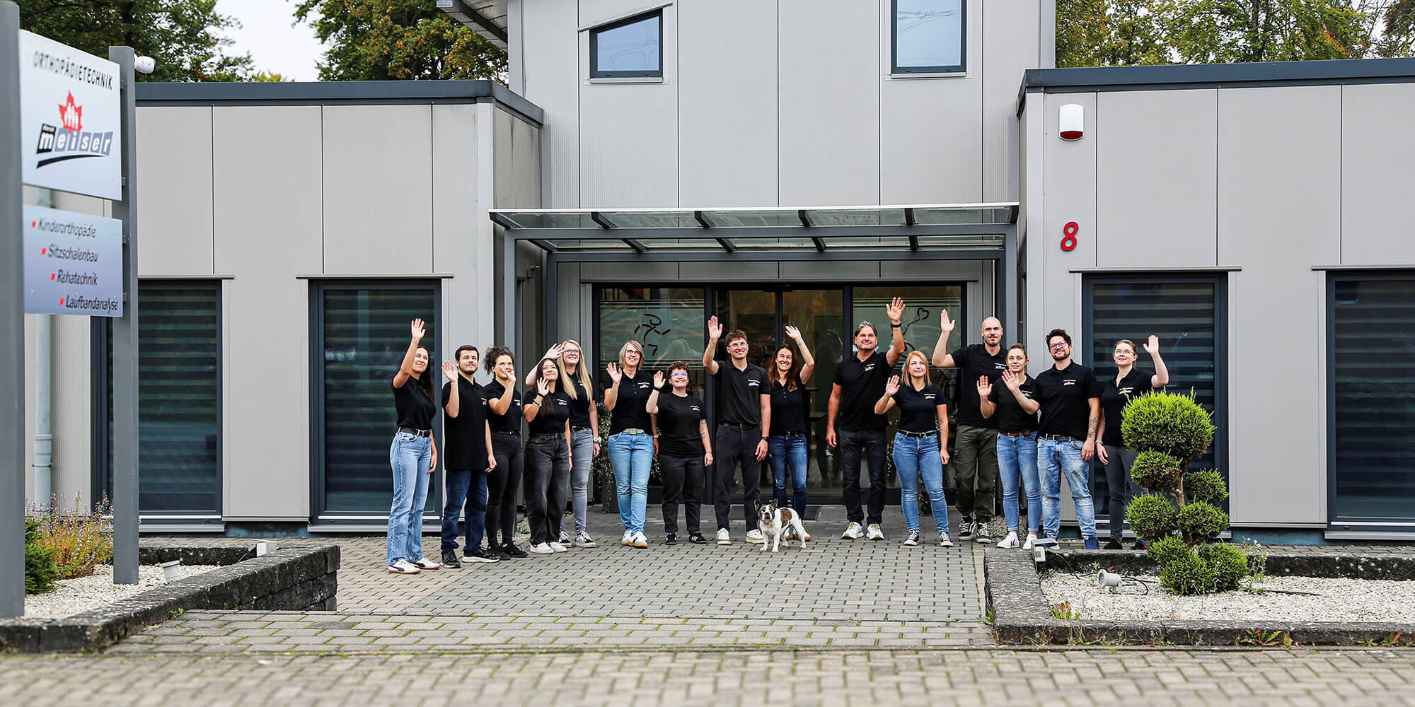Team der Orthopädietechnik steht gemeinsam vor dem Firmengebäude und winkt